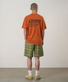 Back view of a model wearing a orange Gramicci G-Sail Tee with black graphic and green plaid pants on a light-tone background.