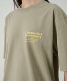 Model wearing a Gramicci beige G-Sail Tee with a yellow graphic against a light-tone background.