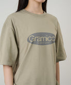 Model wearing a Gramicci beige Outdoor Gear Tee with a grey graphic against a light-tone background.