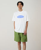 Model wearing a white Gramicci Outdoor Gear Tee with a blue graphic and green shorts on a light-tone background.