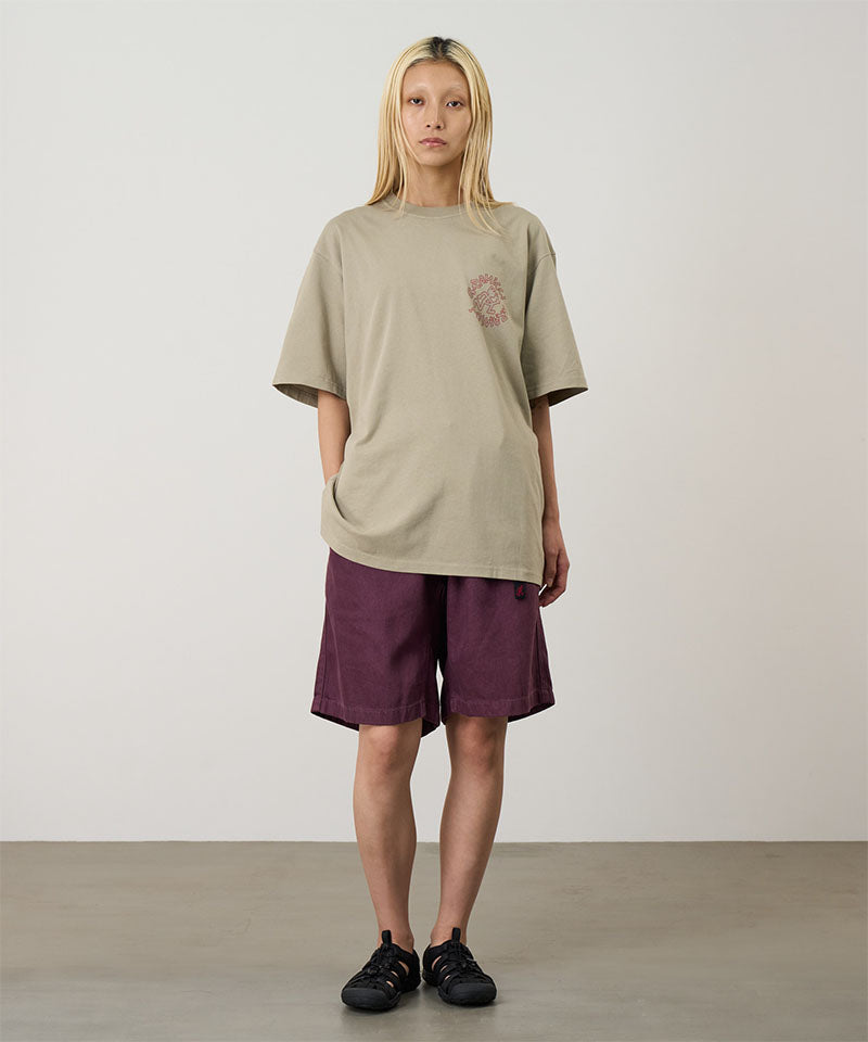 Model wearing a beige Gramicci Running Man Tee with graphic and purple shorts on a light tone background.

