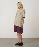 Model wearing a beige Gramicci Running Man Tee with graphic and purple shorts on a light tone background.
