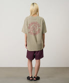 Rear view of a model wearing a beige Gramicci Running Man Tee with graphic with purple shorts on a light tone background.
