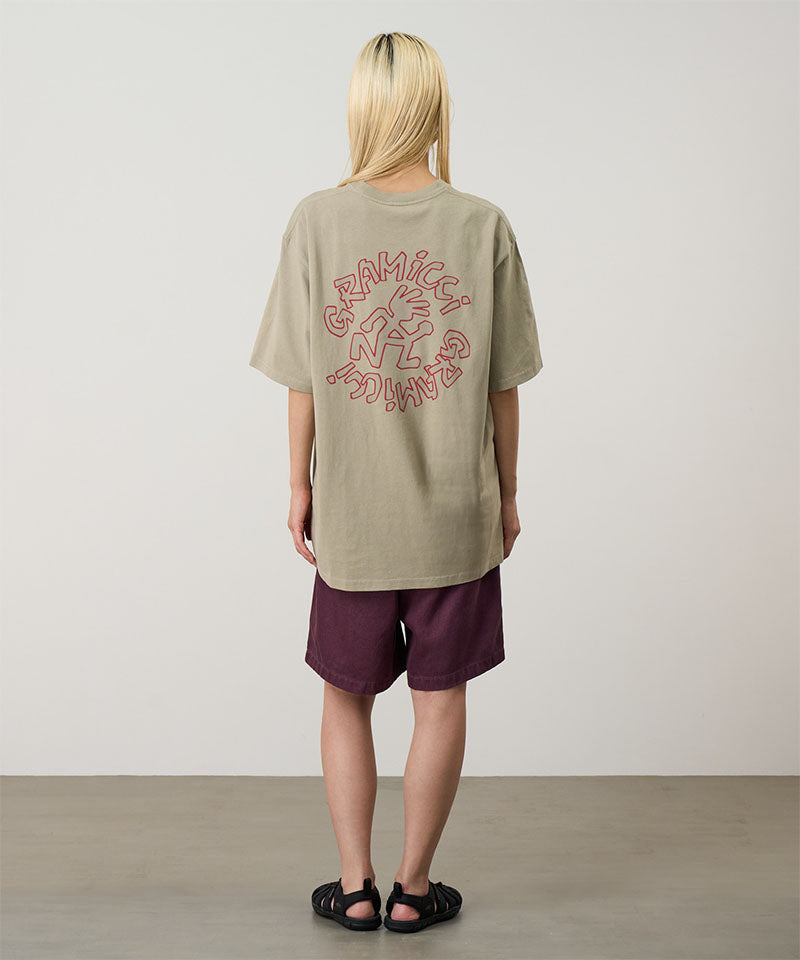 Rear view of a model wearing a beige Gramicci Running Man Tee with graphic with purple shorts on a light tone background.

