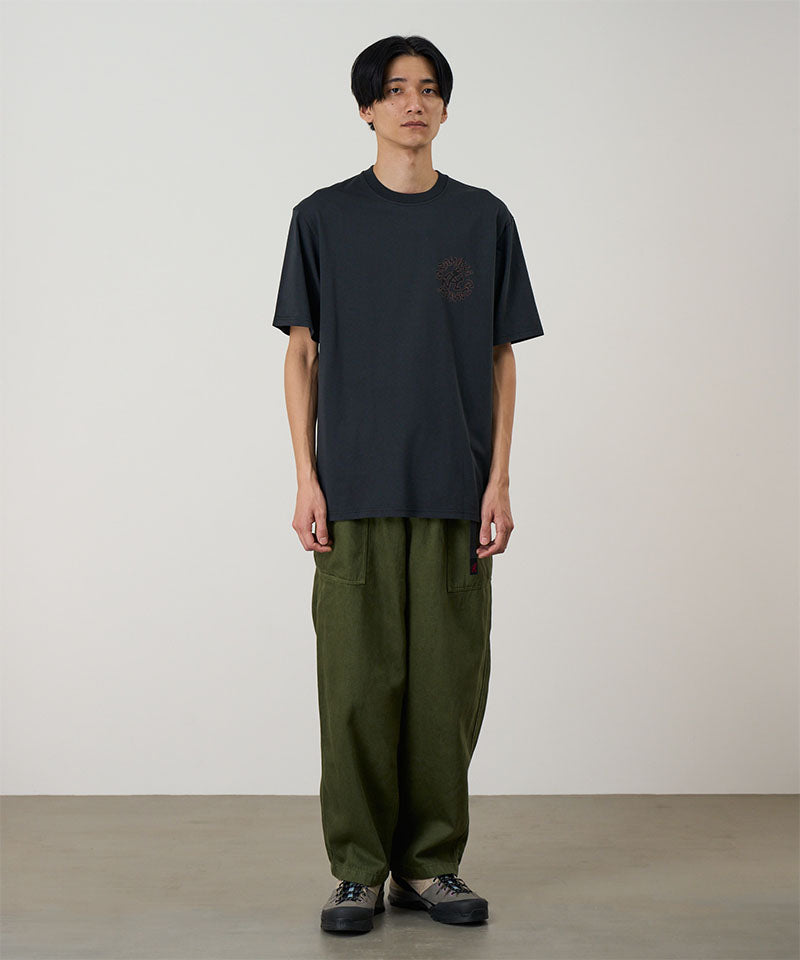 Model wearing a black Gramicci Running Man Tee with graphic and purple shorts on a light tone background.
