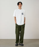 Model wearing a white Gramicci Running Man Tee with graphic and purple shorts on a light tone background.
