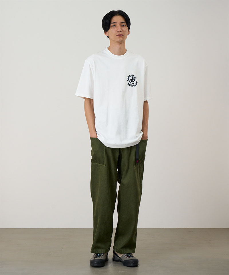 Model wearing a white Gramicci Running Man Tee with graphic and purple shorts on a light tone background.
