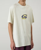 Model wearing a Gramicci natural colored G-Scribble Tee Pigment Dyed shirt with a purple graphic against a light tone background.
