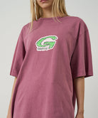 Model wearing a Gramicci red G-Scribble Tee Pigment Dyed shirt with a green graphic against a light tone background.
