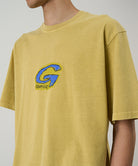 Model wearing a Gramicci sulpher G-Scribble Tee Pigment Dyed shirt with a blue graphic against a light tone background.
