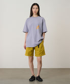 Model wearing a lavender Gramicci Running Man Tee with dark mango colored graphic and yellow shorts on a light-tone background.