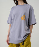 Model wearing a Gramicci lavender Running Man Tee with dark mango colored graphic against a light-tone background.