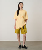 Model wearing a mango colored Gramicci Running Man Tee with dark mango colored graphic and yellow shorts on a light-tone background.