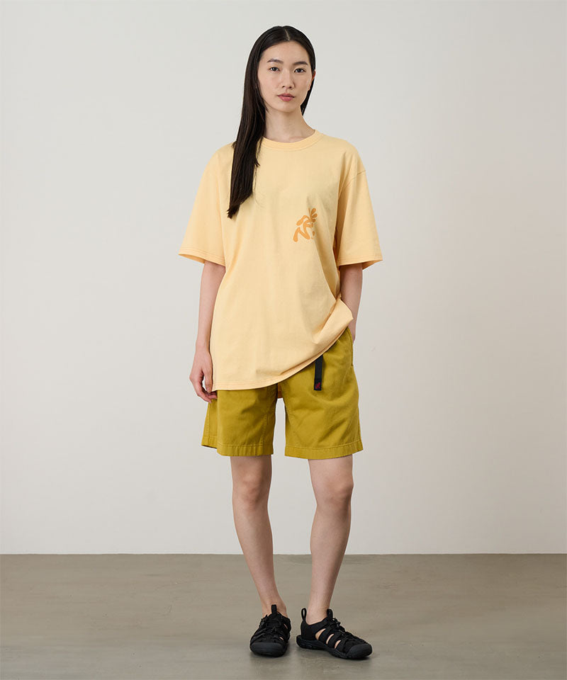 Model wearing a mango colored Gramicci Running Man Tee with dark mango colored graphic and yellow shorts on a light-tone background.