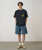 Model wearing a vintage black Gramicci Running Man Tee with lime green graphic and blue plaid shorts on a light-tone background.