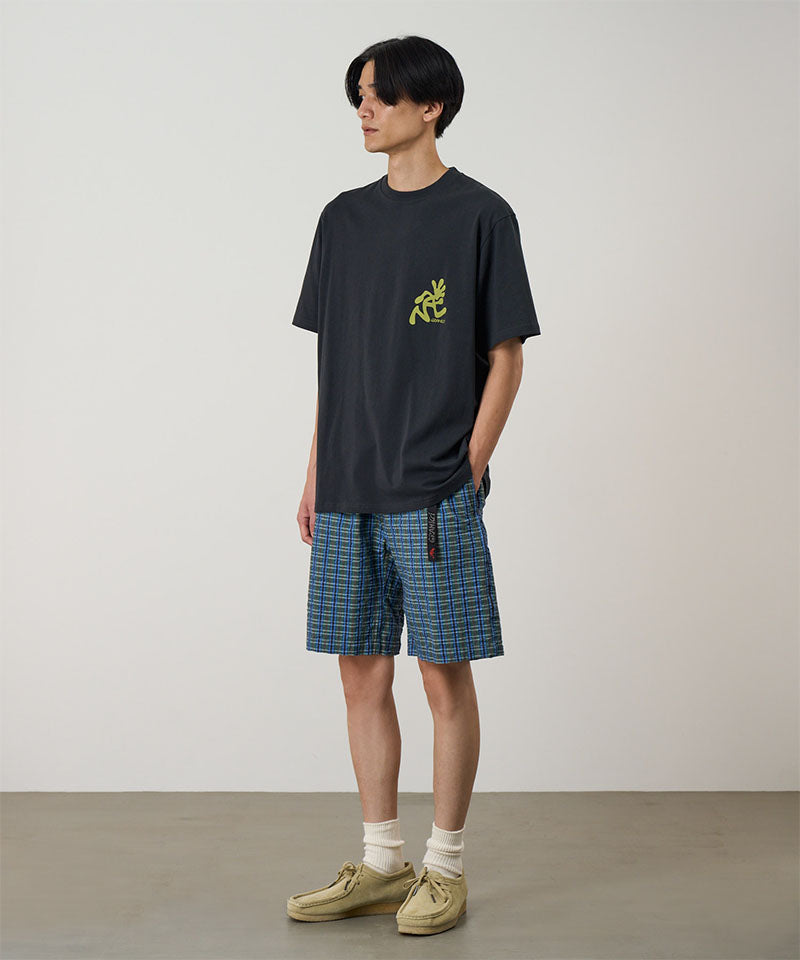 Model wearing a vintage black Gramicci Running Man Tee with lime green graphic and blue plaid shorts on a light-tone background.