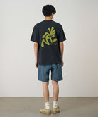 Back view of a model wearing a vintage black Gramicci Running Man Tee with lime green graphic and blue plaid shorts on a light-tone background.