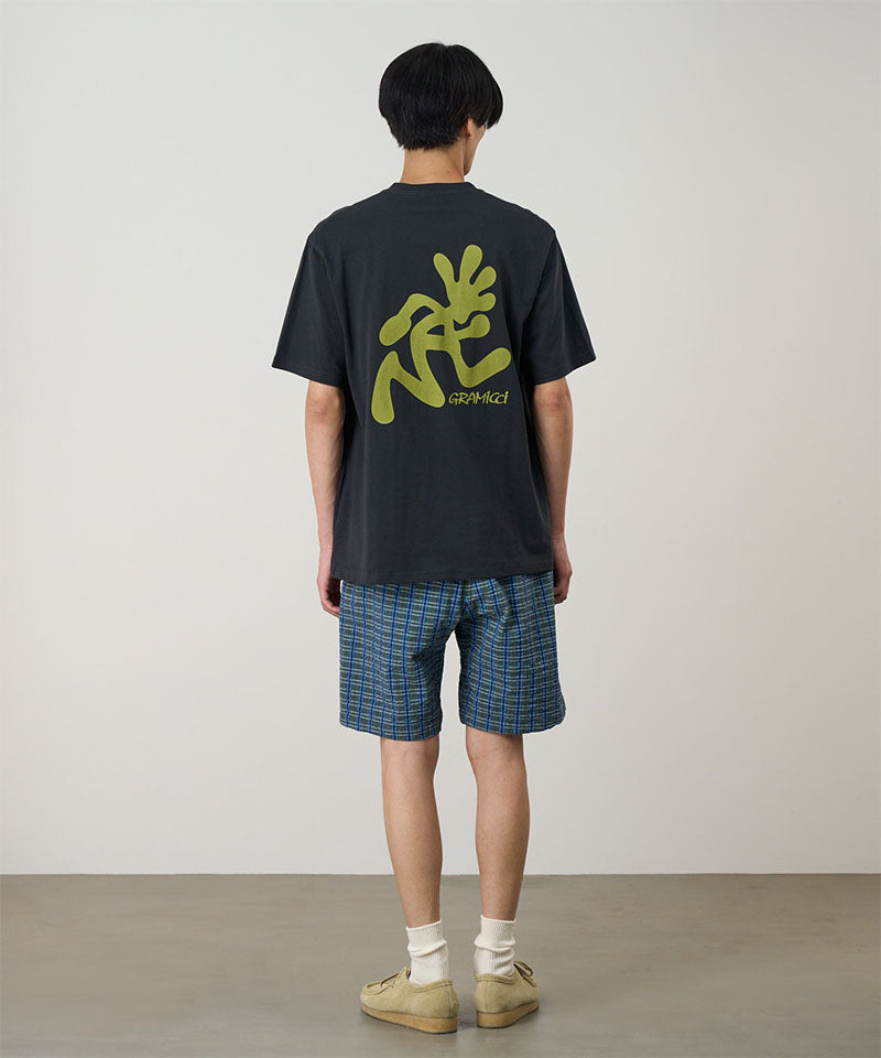 Back view of a model wearing a vintage black Gramicci Running Man Tee with lime green graphic and blue plaid shorts on a light-tone background.