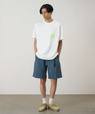 Model wearing a white Gramicci Running Man Tee with lime green graphic and blue plaid shorts on a light-tone background.