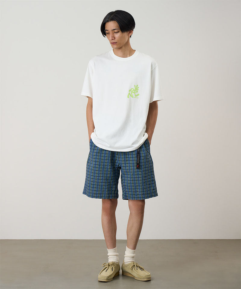 Model wearing a white Gramicci Running Man Tee with lime green graphic and blue plaid shorts on a light-tone background.