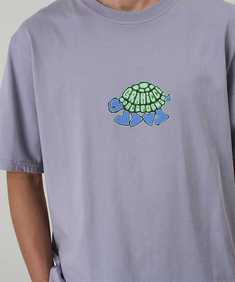 Model wearing a Gramicci lavender Turtle Tee with a turtle graphic against a light tone background.
