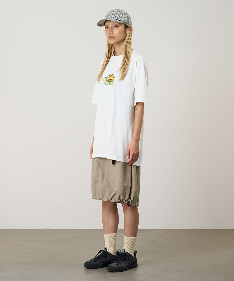 Model wearing a white Gramicci Turtle Tee with a a turtle graphic and beige skirt on a light tone background.
