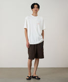 Model wearing a white Gramicci Ice Sculpture Tee with a Gramicci graphic and blue shorts on a light tone background.