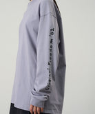 Model wearing a Gramicci lavender Freedom L/S Tee with black graphic on sleeves against a light tone background.
