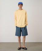 Model wearing a mango colored Gramicci Freedom L/S Tee with a dark mango colored graphic on sleeves and blue checkered shorts on a light tone background.
