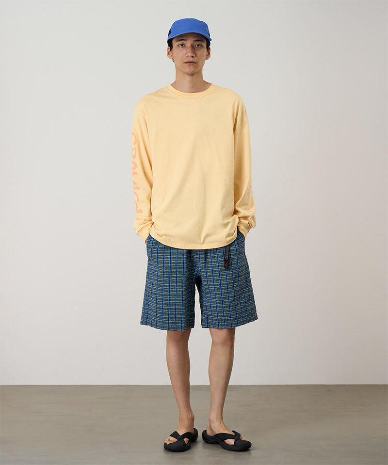 Model wearing a mango colored Gramicci Freedom L/S Tee with a dark mango colored graphic on sleeves and blue checkered shorts on a light tone background.
