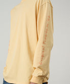 Model wearing a Gramicci mango colored Freedom L/S Tee with dark mango colored graphic on sleeves against a light tone background.
