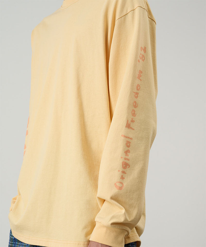 Model wearing a Gramicci mango colored Freedom L/S Tee with dark mango colored graphic on sleeves against a light tone background.
