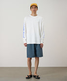 Model wearing a white Gramicci Freedom L/S Tee with a blue graphic on sleeves and blue checkered shorts on a light tone background.
