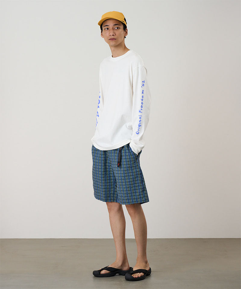 Model wearing a white Gramicci Freedom L/S Tee with a blue graphic on sleeves and blue checkered shorts on a light tone background.
