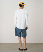 Back view of a model wearing a white Gramicci Freedom L/S Tee with a blue graphic on sleeves and blue checkered shorts on a light tone background.
