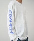 Model wearing a Gramicci white Freedom L/S Tee with blue graphic on sleeves against a light tone background.
