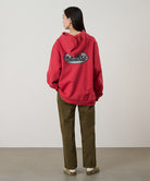 Back view of a model wearing bark pigment colored Gramicci Women's Brooks Pants and a red hoodie on a light-tone background.