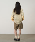Back view of a model wearing beige Gramicci Women's Corduroy Utility Short and a green plaid shirt on a light-tone background.