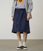 Model wearing a british anavy colored Gramicci Nylon Packable Midi Skirt and plaid shirt over a sunflower tee on a light-tone background.