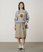 Model wearing a milk tea colored Gramicci Nylon Packable Midi Skirt and plaid shirt over a sunflower tee on a light-tone background.