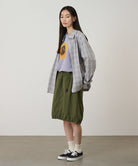 Model wearing an olive colored Gramicci Nylon Packable Midi Skirt and plaid shirt over a sunflower tee on a light-tone background.