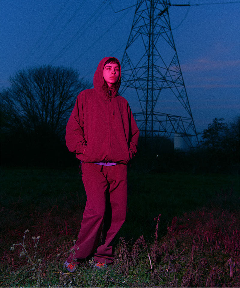 Woman wearing a red Gramicci jacket and pants standing in a field with power lines in the background