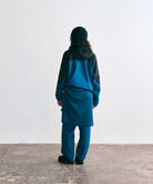 Model wearing a Gramicci teal and black colorblock hooded jacket, matching teal pants with a sweatshirt tied around the waist, a black beanie, and black trail shoes, seen from behind.   