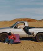 Two people relaxing on a pink cushion in front of a white pickup truck in a desert setting.