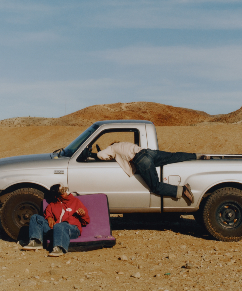 Two people relaxing on a pink cushion in front of a white pickup truck in a desert setting.