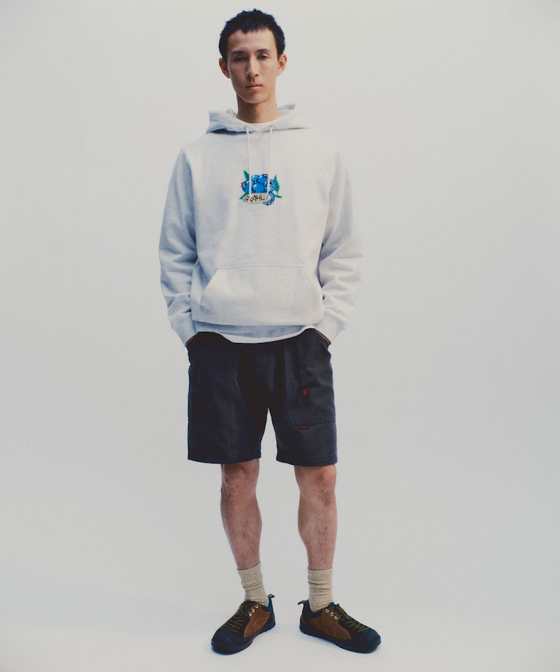 Model wearing the Gramicci Blueberry Hooded Sweatshirt with blueberry graphic in heather ash and navy colored shorts on a light tone background.
