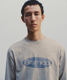 Model wearing a beige Gramicci Outdoor Gear L/S Tee with a blue graphic against a light-tone background.