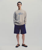 Model wearing a beige Gramicci Outdoor Gear L/S Tee with a blue graphic and blue shorts against a light-tone background.