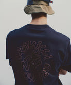 Rear view of a model wearing a navy Gramicci Running Man Tee with graphic against a light tone background.

