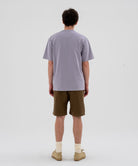 Back view of a model wearing a lavender Gramicci Half Dome Tee and brown shorts on a light-tone background.
